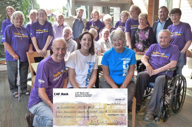 Cheque presentation by Lynne Turner of Vocal Fusion with Timothy Medhurst from Dukes of Dorchester ‘celebrating’ in the background.